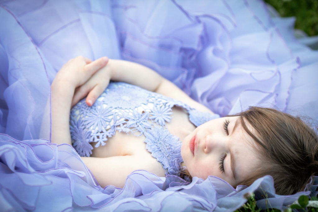 Young girl with brown hair and long eyelashes sleeping in a field with her hands folded over her stomach.  She is wearing a periwinkle blue dress from Butterfly Closet.
