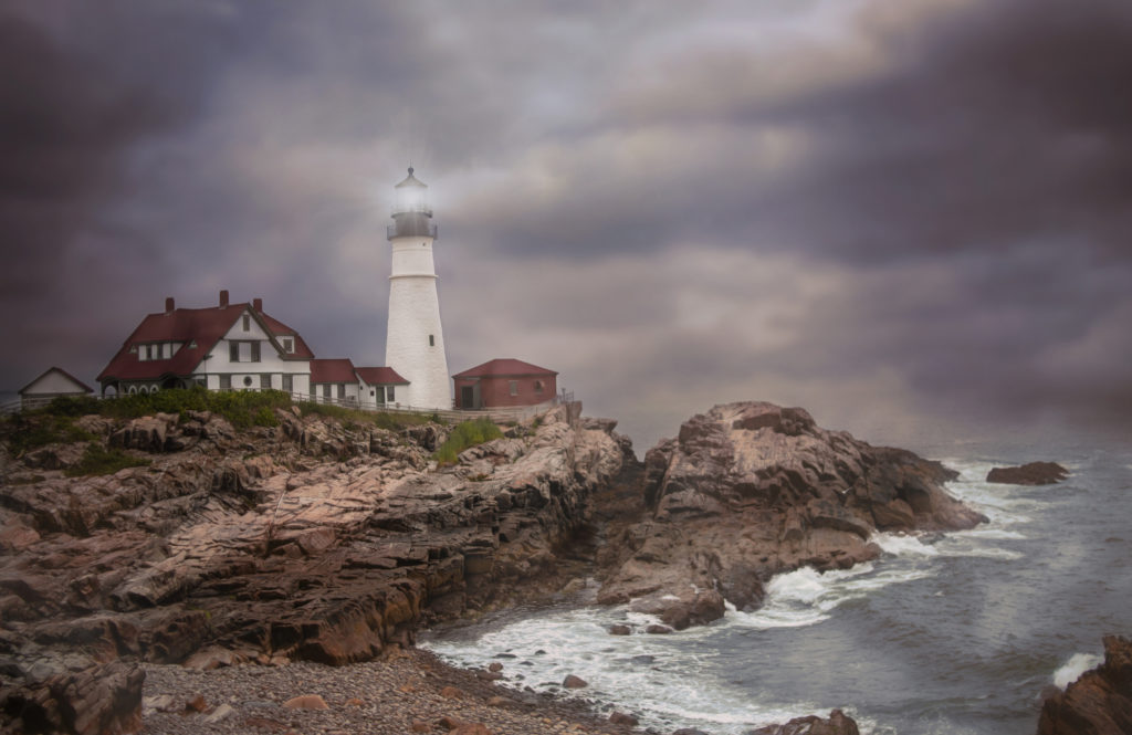 The Portland Head Light in Portland, Maine on a stormy day.  There are dark clouds in the background and waves crashing on to the rocky shore.  The lighthouse is white.