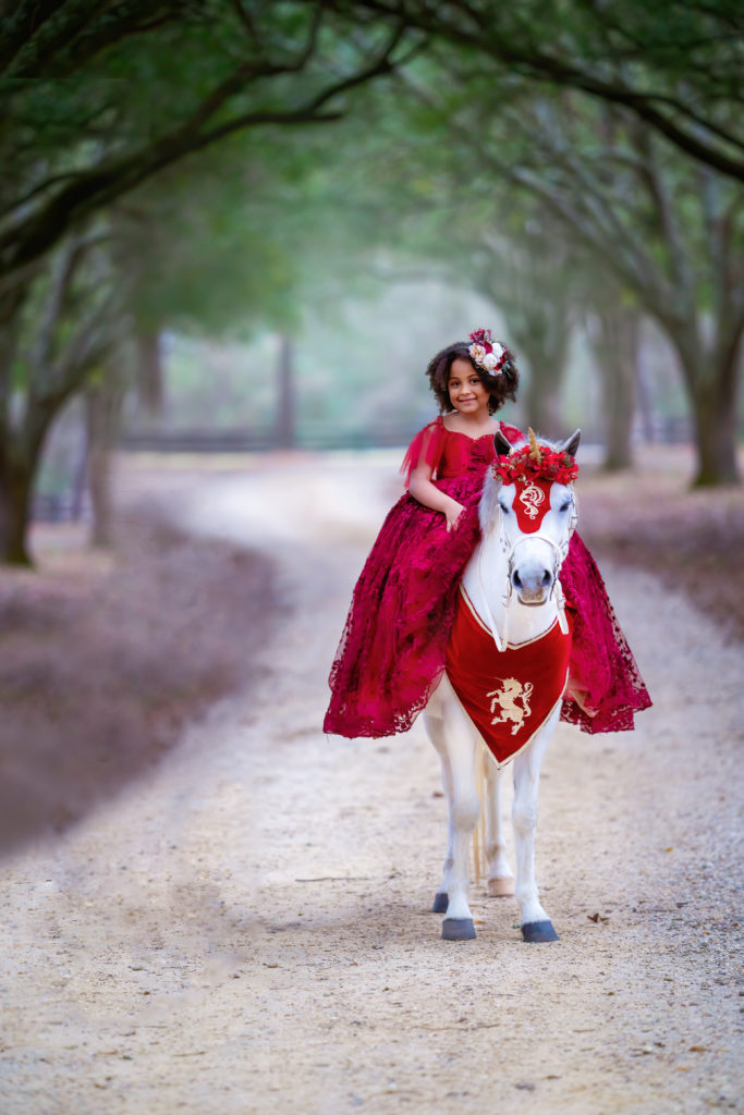 Black girl on a white welsh pony dressed in a red gown from Bentley & Lace under an oak alley in Covington Louisiana