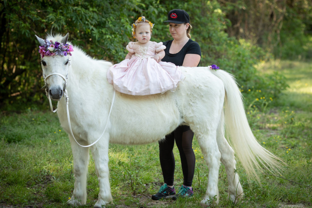 Behind the Scenes image of a mom holding her baby daughter on the back of a white welsh pony that has a floral headband and unicorn horn. Mom is safely holding the baby for her unicorn photo session.
