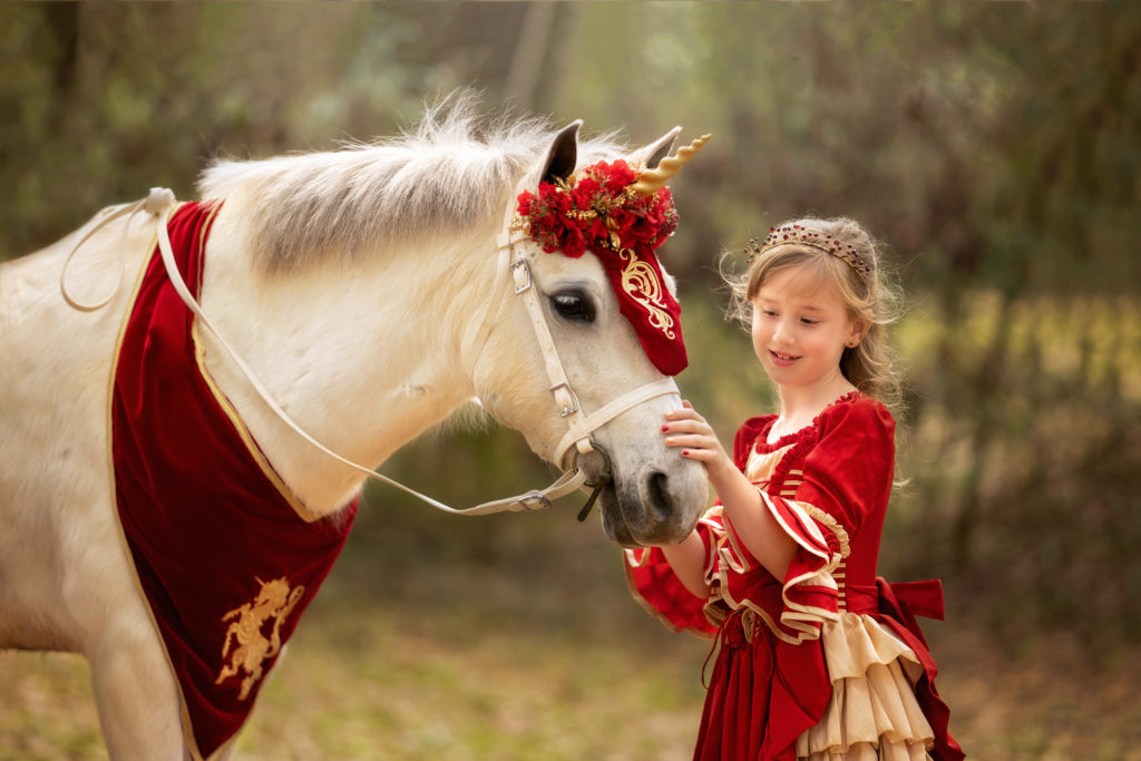 7 year old girl wearing a red princess dress petting the nose of a white Welsh pony with a gold unicorn horn, red bridle and floral headband, and red embroidered scarf.  They are standing in the fall leaves in a New Orleans, Louisiana.