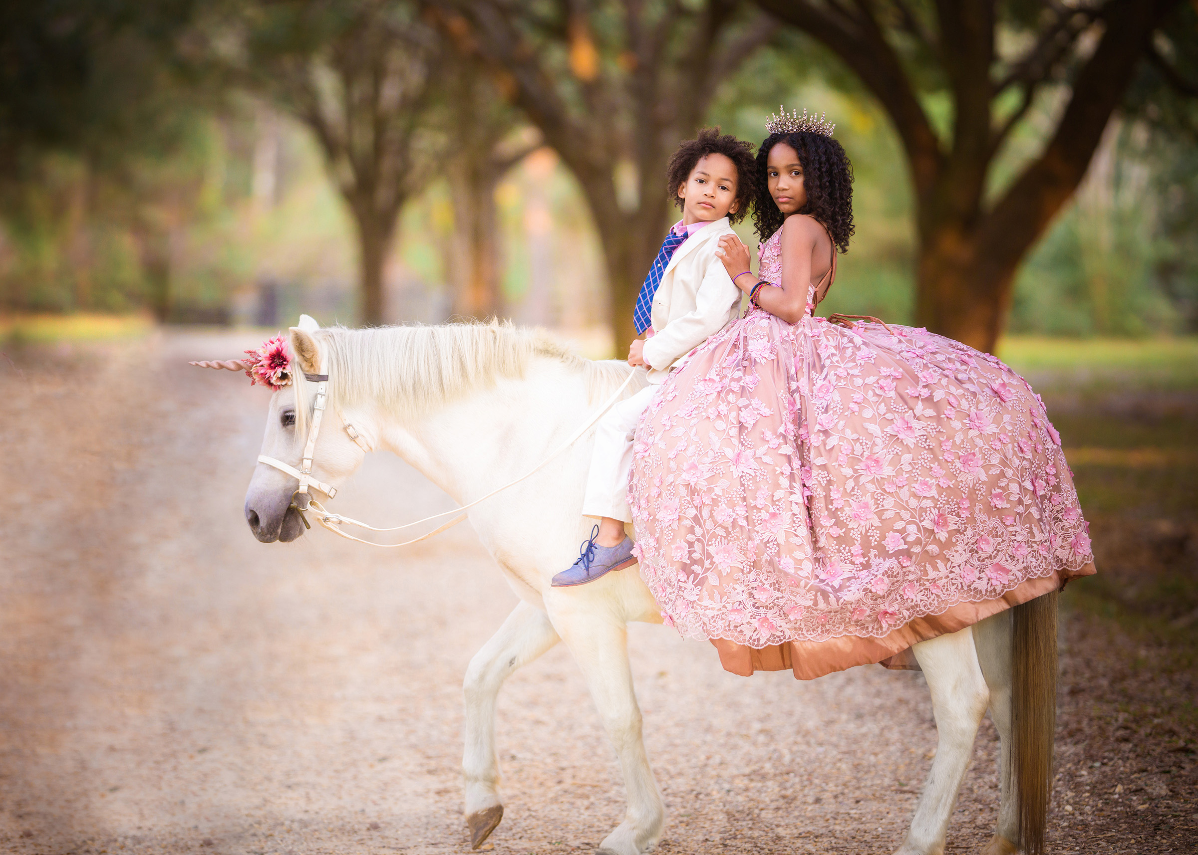 Photo of black brother and sister on a white unicorn riding the horse on a gravel path under oak trees