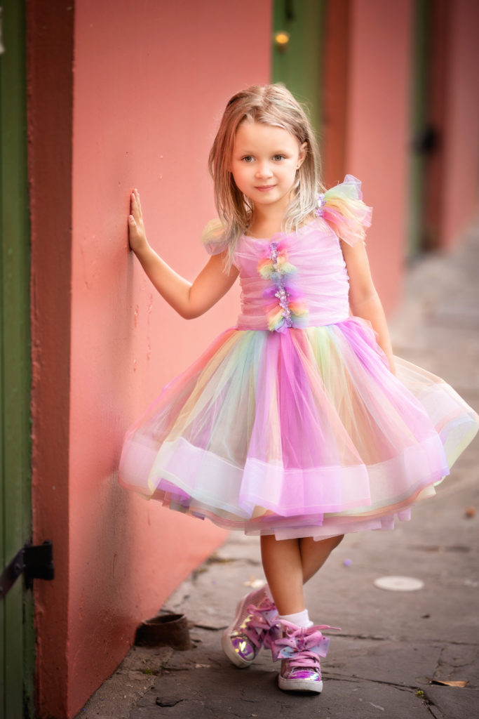 Little girl with blond hair wearing a poufy rainbow dress and shiny high tops.  She is leaning against an orange building in the French Quarter.