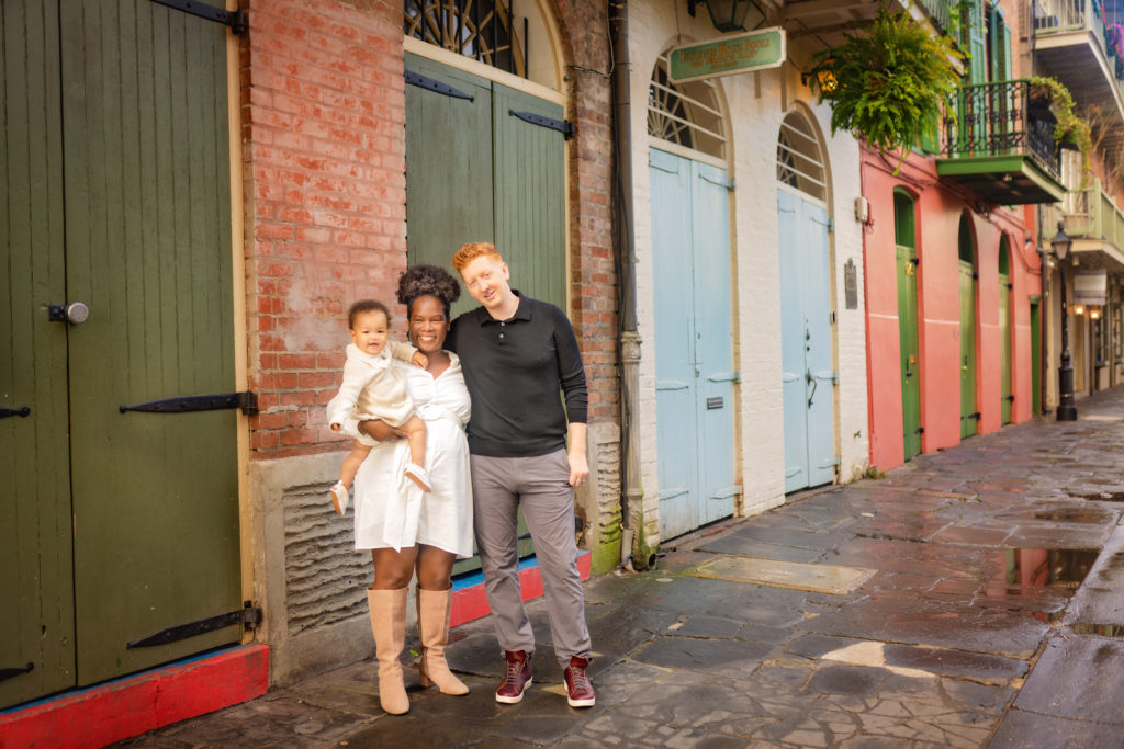 Black woman and white man  with red hair standing in front of a brick building in the French Quarter.  The building has green shutters and the couple is holding a baby with curly black hair.  The woman is wearing a white dress and brown boots, and the man is wearing a black shirt and gray pants. 