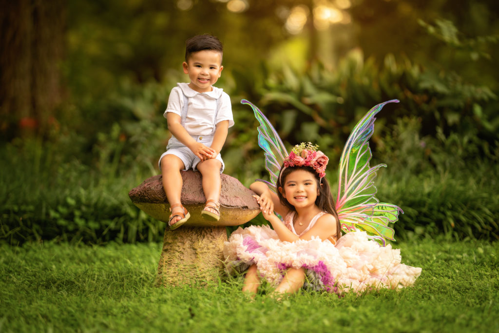 Asian brother and sister in sitting in the grass in Audubon Park.  The girl is wearing fairy wings and smiling.  The boy is sitting on top of a mushroom.  