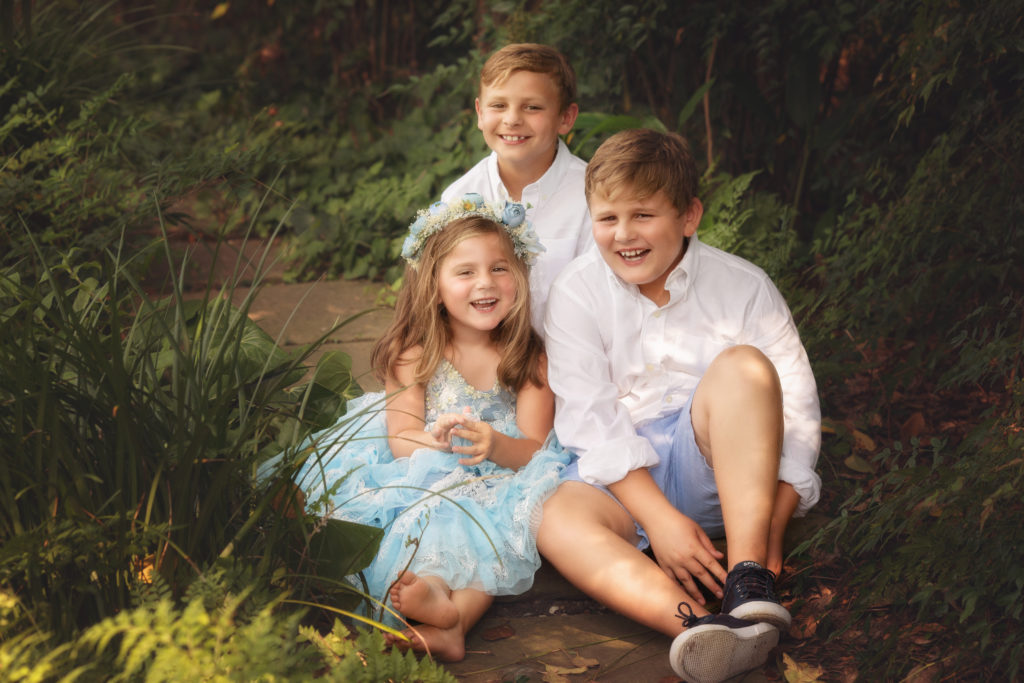 Two boys and a girl and sitting on the ground in between plants giggling and laughing.  The boys are both wearing white shirts and blue shorts.  The girl is the youngest and is wearing a flower crown and pale blue dress with white lace.