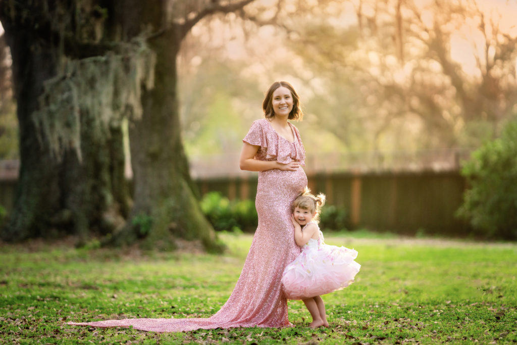Pregnant mom with short brown hair is standing in the park under the oak trees.  She is wearing a pink, sequin dress that shows off her belly.  Her toddler girl is hugging her legs and smiling.