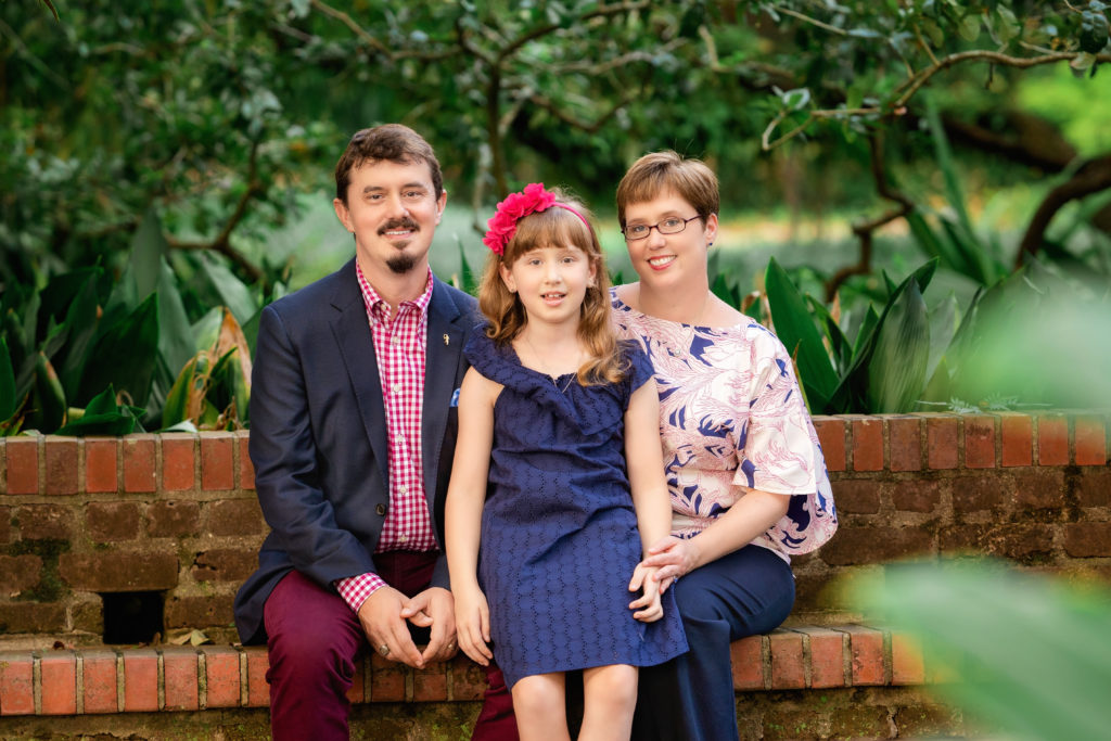 Man, woman, and child sitting on a brick bench in City Park.  The girl is wearing a navy dress and a pink floral headbank.  The mom is wearing a print shirt and navy pants.  The man has a moustache and goatee and is wearing a burgundy gingham shirt, navy blazer, and burgundy slacks.