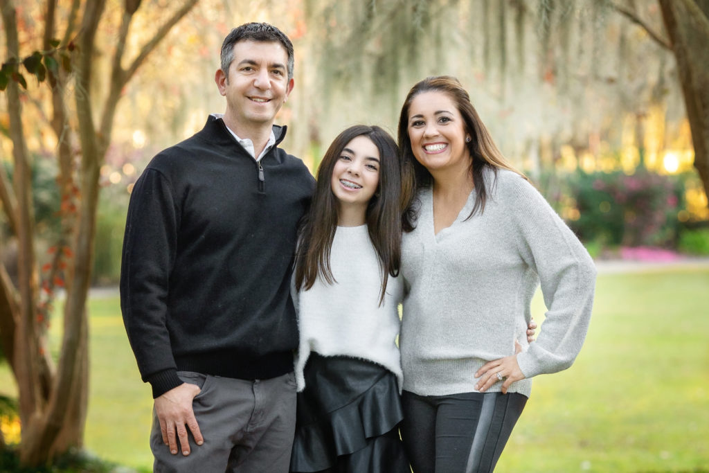 A family smiling under the Spanish Moss in Audubon Park.  The dad has graying hair, the mom has long, brown hair, and the teenage daughter has long brown hair.  They are all smiling and looking at the camera.