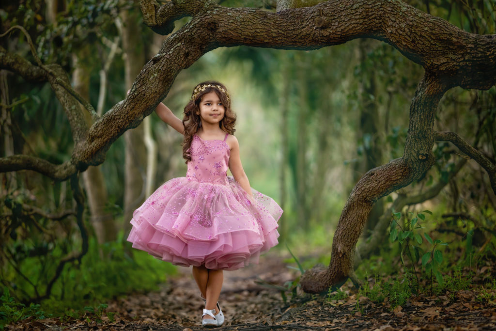 5 year old girl with long, curly brown hair and olive skin, wearing a mauve star dress and holding onto an oak limb in City Park.