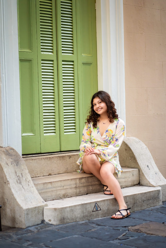Hispanic female teenager sitting on some steps in front of a green shuttered door in the French Quarter.  She has brown curly hair and is wearing a floral romper.  She is smiling. 