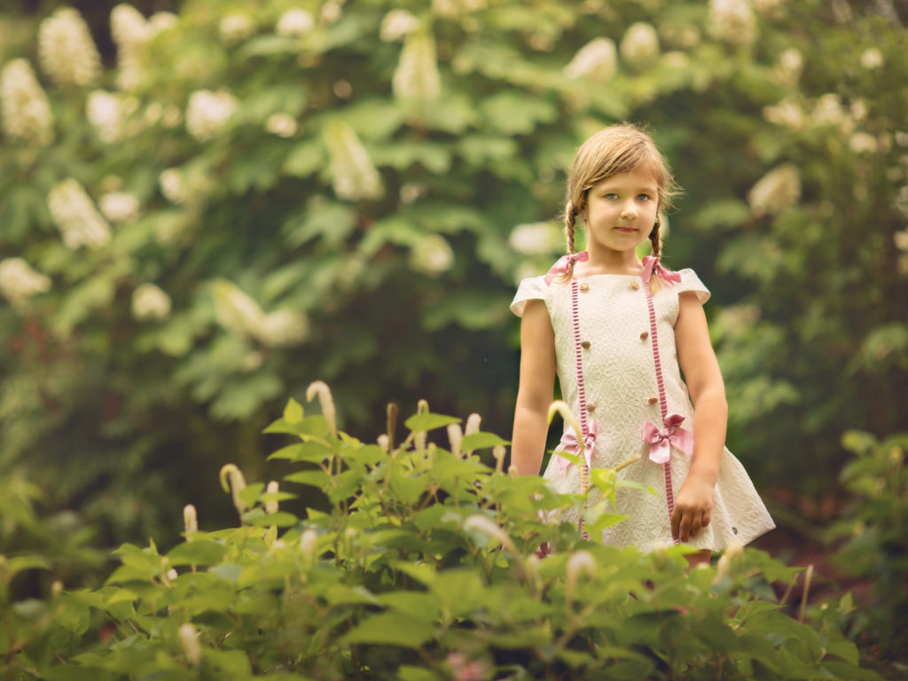 A small blonde girl in a vintage style white dress is standing among white blooming flowers in the gardens.  She has two braids and is looking at the camera.  Her dress is white with pink bows.