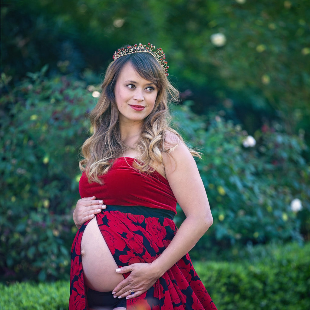 Hispanic woman who is 8 months pregnant is wearing a red and black floral and velvet dress.  She is also wearing a red jeweled crown.  She has long, dark curly hair and is looking over her shoulder.  Her belly is exposed.