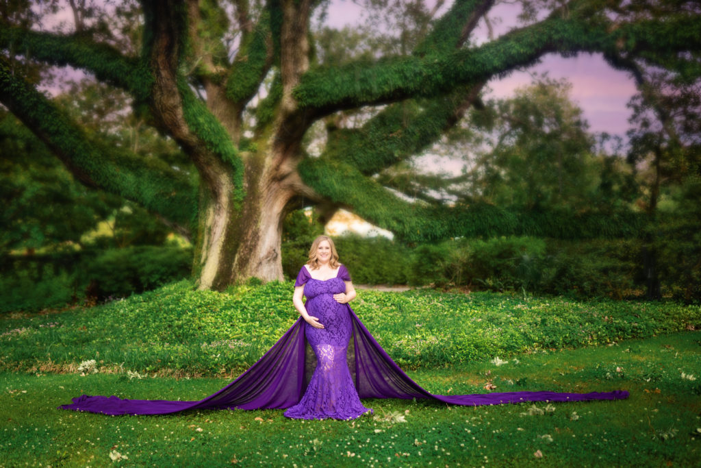 Maternity photo of a woman with shoulder length light brown hair, wearing a purple mermaid style dress and a very long train.  She is holding her belly and standing under a mossy oak tree.  The sun is setting and the sky is lavender.