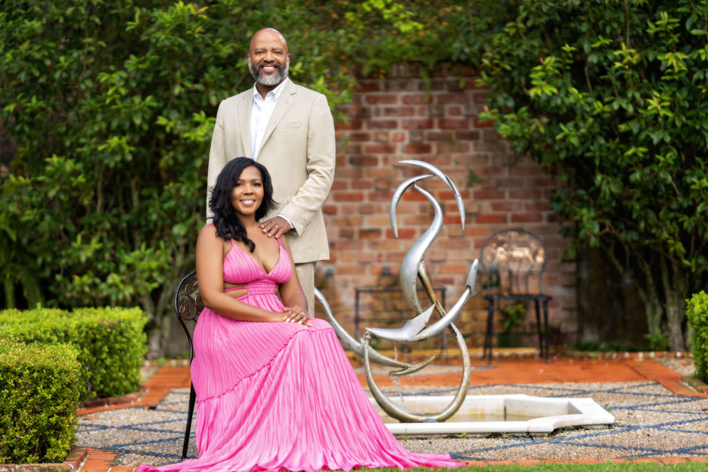 Beautiful black man and woman dressed formally, posing in front of a brick wall in Longue Vue gardens.  The woman is wearing a pink flowy dress and is sitting on a chair.  The man has his arm on her shoulder and is wearing a tan linen suit.